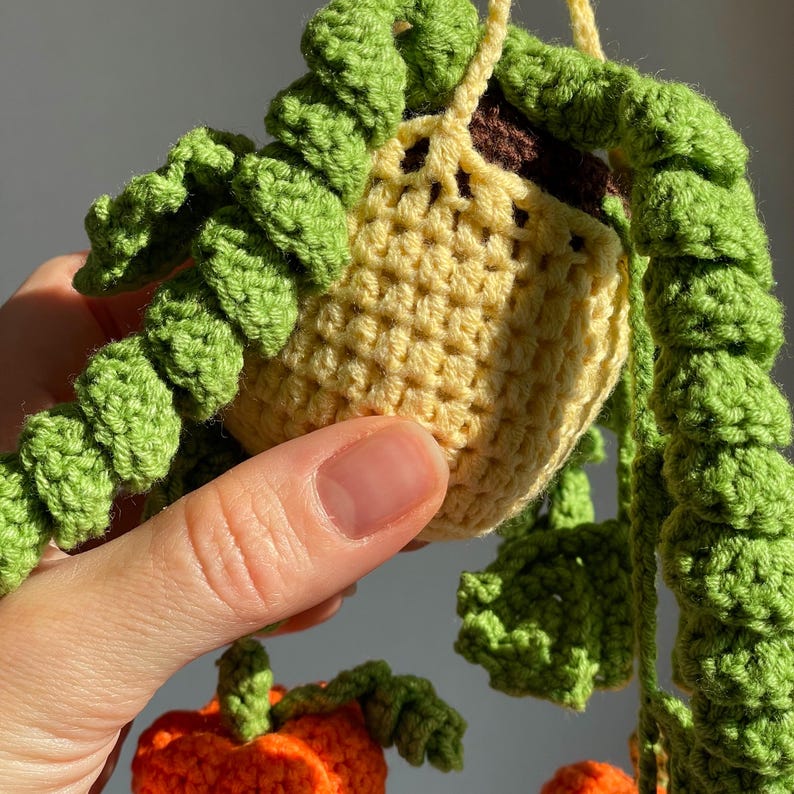 May include: A crocheted hanging plant in a light yellow pot with dark brown soil, featuring long, trailing green vines with textured leaves. Below the pot, a small orange crocheted pumpkin with green leaves is visible, all crafted from yarn.