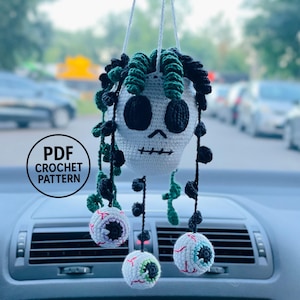 May include: A white crocheted skull hanging ornament with dark green and black curly vines. Three small crocheted eyeballs with red veins hang from the skull by black strings. A circular black logo with the text PDF CROCHET PATTERN is visible.