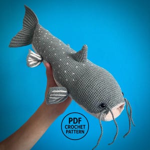 May include: A gray crocheted catfish toy featuring a textured surface with small white speckles, a light-colored underside, dark eyes, and long whiskers. A circular black and white logo on the image contains the text PDF CROCHET PATTERN.