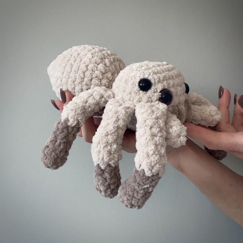May include: A plush, crocheted spider toy in cream and gray. The spider has large, black eyes and eight legs. The legs are cream-colored with gray tips. The body is a soft, textured material.