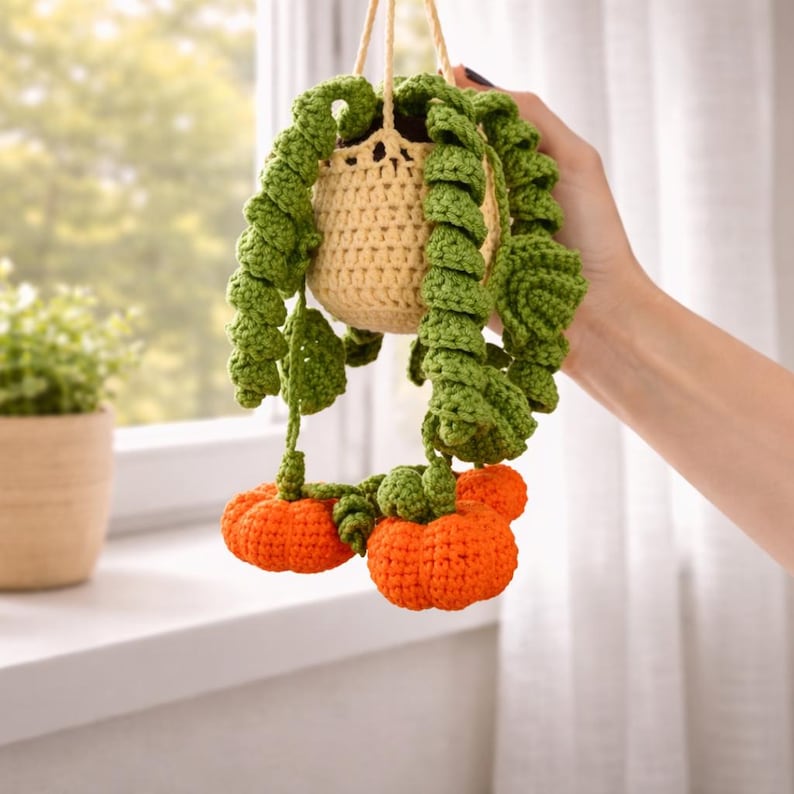 May include: A crocheted hanging plant featuring a light beige pot, long spiraling green vines, and three small orange pumpkins attached to the ends of the vines. The entire piece is crafted from yarn with visible stitch textures throughout the design.