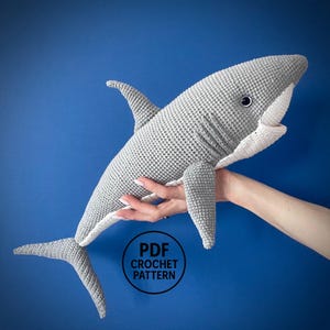 May include: A gray and white crocheted shark toy featuring a dark eye, a dorsal fin, pectoral fins, and a tail. A circular black logo with the white text PDF CROCHET PATTERN is positioned in the lower center of the image against a blue background.
