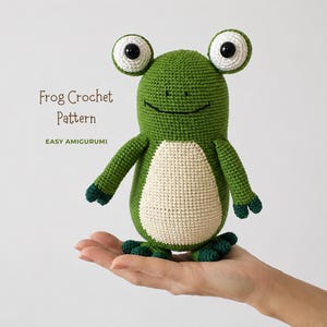 May include: A green and cream crocheted frog amigurumi toy with large white eyes and black pupils. The text 'Frog Crochet Pattern' and 'EASY AMIGURUMI' is printed in brown and green on the left side of the green crocheted frog figure.