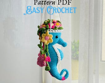 May include: A crocheted blue seahorse with a white trim and a flower pot on its head. The pot is filled with pink, yellow, and green crocheted flowers. The seahorse is hanging from a white string. The text "Pattern PDF EASY CROCHET" is visible in the image.