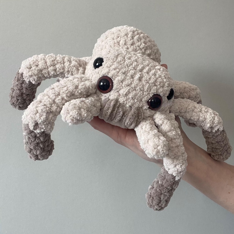 May include: A handmade, plush spider toy in cream and gray. The spider has eight legs, large brown eyes, and a textured, fuzzy body. The tips of the legs are a darker gray. The toy is held in a person's hand.