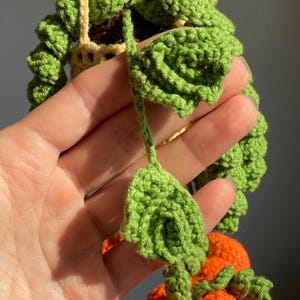 May include: A crocheted hanging plant featuring vibrant green leaves and small orange spherical elements. The plant is suspended by a light yellow crocheted cord, with detailed green foliage cascading downwards in a textured, handmade yarn construction.