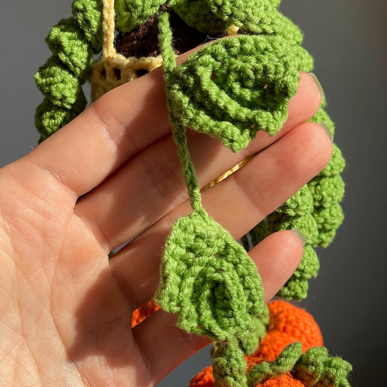 May include: A crocheted hanging plant featuring vibrant green leaves and small orange spherical elements. The plant is suspended by a light yellow crocheted cord, with detailed green foliage cascading downwards in a textured, handmade yarn construction.