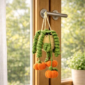 May include: A crocheted hanging plant decoration featuring a light yellow pot with long, spiraling green vines and three small, round, textured orange pumpkins dangling from the ends of the vines, suspended by a light yellow yarn cord.