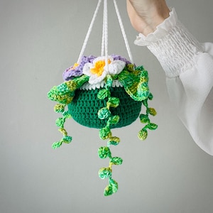 May include: A handmade crochet hanging planter in shades of green, purple, white, and yellow. The planter features a green pot with a crocheted flower and trailing vines. The planter is suspended by white cords.