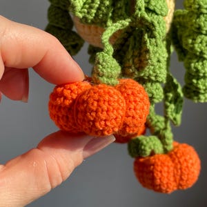 May include: A crocheted hanging plant decoration featuring bright orange pumpkins with textured, rounded segments and vibrant green crocheted leaves and vines. The small, handcrafted pumpkins are attached to green stems that hang downward from a central point.