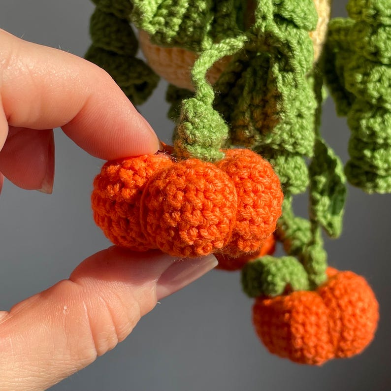 May include: A crocheted hanging plant decoration featuring bright orange pumpkins with textured, rounded segments and vibrant green crocheted leaves and vines. The small, handcrafted pumpkins are attached to green stems that hang downward from a central point.