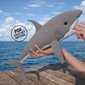 Könnte beinhalten: Ein gehäkelter Hai in Grau mit weißen Akzenten, einschließlich Maul und Unterseite. Der Hai hat eine Rückenflosse, Brustflossen und eine Schwanzflosse. Das Bild enthält eine kreisförmige Grafik mit der Aufschrift "PDF Crochet Pattern".