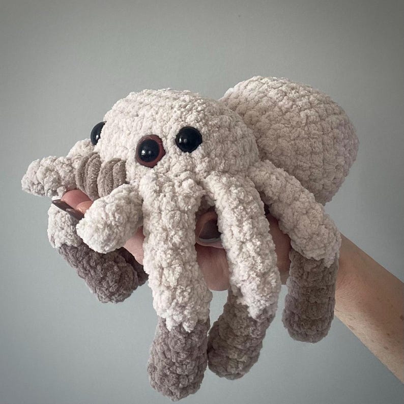 May include: A plush, crocheted spider toy in cream and taupe. The spider has large, black eyes with brown centers and eight legs. The legs and body are made of a soft, textured yarn. The toy is held in a hand.