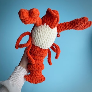 May include: A plush, orange lobster toy with a white belly. The lobster has large claws and a segmented tail. The toy is made of a soft, textured material, and is held against a light blue background.