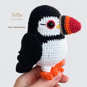 Może przedstawiać: Szydełkowana zabawka amigurumi przedstawiająca maskonura wykonana z czarnej, białej i pomarańczowej włóczki. Ptak ma czarną głowę i skrzydła, białą twarz i pierś oraz jaskrawopomarańczowy dziób i stopy. Tekst na obrazku brzmi Puffin Crochet pattern EASY AMIGURUMI.