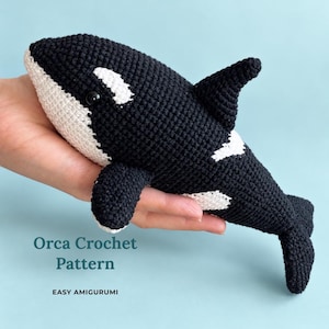 Puede incluir: Un juguete de orca tejido a ganchillo en blanco y negro con cabeza redondeada, aleta dorsal y aletas laterales. El texto "Orca Crochet Pattern" y "EASY AMIGURUMI" está impreso en azul verdoso oscuro en la parte inferior.