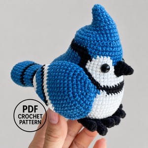 May include: A blue jay bird amigurumi crochet toy featuring a bright blue head and back, a white chest and face, and black accents around the eyes and wings. The bird has a small black beak and black feet. A circular logo reads PDF CROCHET PATTERN.