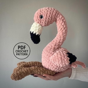 May include: A handmade pink crochet flamingo with a white and black beak. The flamingo has a long, curved neck and is perched on a brown crocheted base. The image includes a circular graphic that says "PDF CROCHET PATTERN".