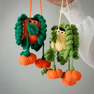 May include: Two hanging crochet plant decorations featuring orange and yellow pots with green leafy vines and small orange pumpkin-shaped accents. Each piece is suspended by a matching yarn cord, showcasing intricate stitch work and vibrant colors.