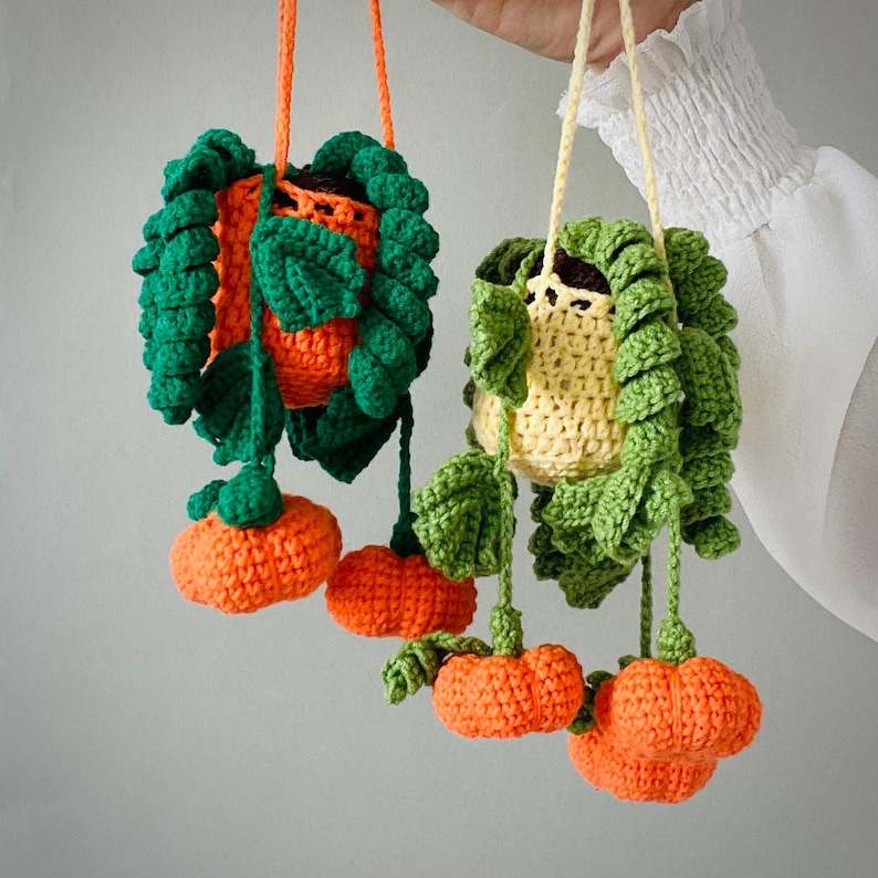 May include: Two hanging crochet plant decorations featuring orange and yellow pots with green leafy vines and small orange pumpkin-shaped accents. Each piece is suspended by a matching yarn cord, showcasing intricate stitch work and vibrant colors.