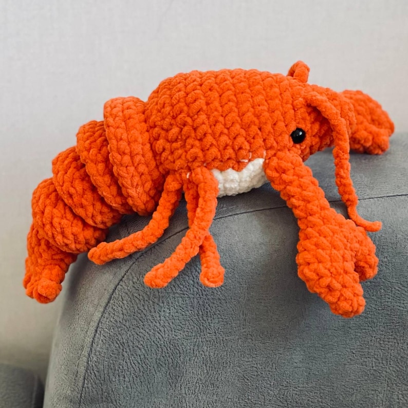 May include: A handmade, plush lobster toy in vibrant orange. The crochet lobster features a segmented body, detailed claws, and small black eyes. The underside of the lobster is white. This soft toy is a unique gift for children or a nautical-themed decor item.