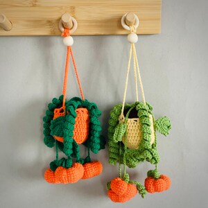 May include: Two crocheted hanging plant baskets featuring orange and yellow pots with green trailing vines and small orange pumpkin accents. Each basket is suspended by a matching yarn cord attached to a wooden bead and a light wood wall hook.