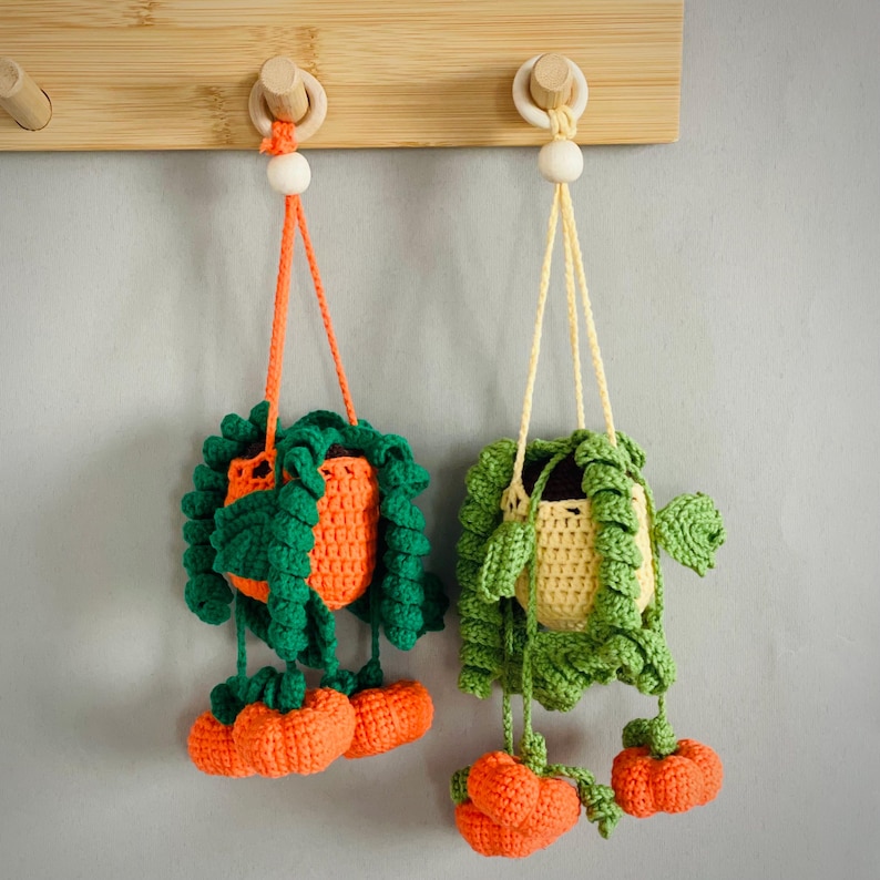 May include: Two crocheted hanging plant baskets featuring orange and yellow pots with green trailing vines and small orange pumpkin accents. Each basket is suspended by a matching yarn cord attached to a wooden bead and a light wood wall hook.