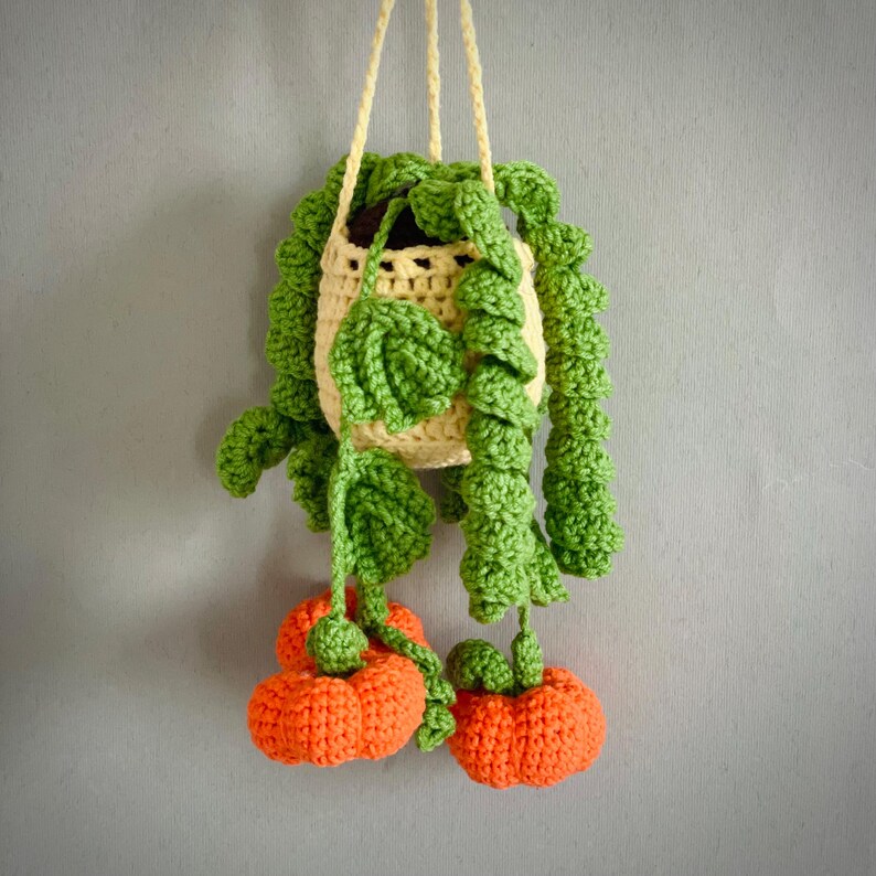 May include: A crocheted hanging plant featuring a cream-colored yarn pot with long, trailing green vines and leaves. Two bright orange, textured, pumpkin-shaped crocheted ornaments hang from the ends of the vines, suspended by yellow yarn.