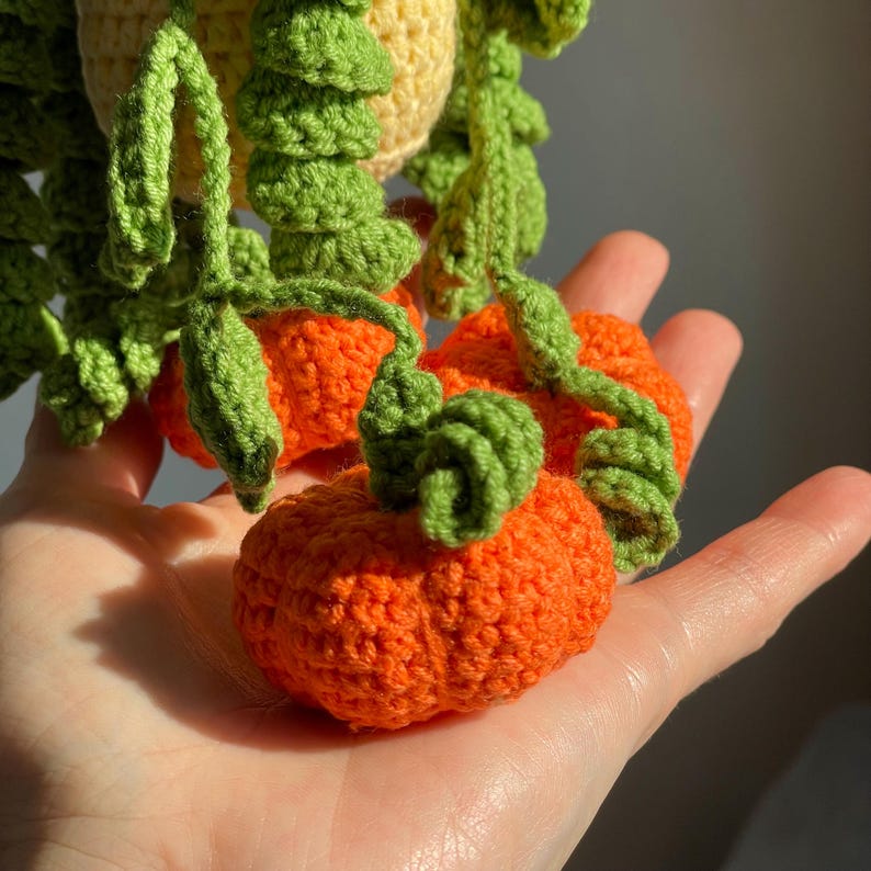 May include: A small, handcrafted crochet decoration featuring bright orange pumpkins with textured green vines and leaves. The pumpkins are attached to a light yellow crocheted base, creating a hanging plant aesthetic with detailed, spiraled green tendrils.