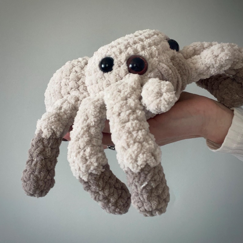 May include: A plush, crocheted spider toy in shades of cream and brown. The spider has large, round, black and brown eyes and eight legs. The legs are two-toned, with cream-colored bodies and brown tips. The toy is soft and cuddly.