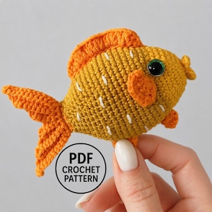 May include: A mustard yellow and orange crocheted fish toy with a single green eye, textured fins, and white stitched accents. A circular black and white label is overlaid on the image with the text PDF CROCHET PATTERN in bold letters.
