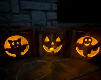 May include: Three wooden Halloween decorations with carved designs. The first is a bat, the second is a jack-o'-lantern, and the third is a ghost. Each decoration has a warm, orange glow from a light inside.