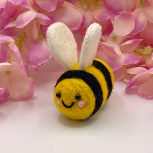 May include: A small, yellow and black felt bee with white wings and a smiling face. The bee is sitting on a white surface with pink flowers in the background.