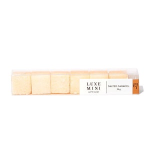 May include: A clear plastic box containing six small, square, light brown sugar cubes. The box has a label that reads "LUXE MINI LATTE CUBE SALTED CARAMEL 24g".