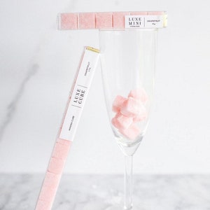May include: A clear plastic tube filled with pink sugar cubes. The tube is labeled "LUXE MINI MIMOSA CUBE GRAPEFRUIT 24g". A glass of champagne with pink sugar cubes in it.