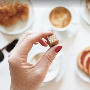 May include: A hand holding a brown sugar cube. The sugar cube is in focus, while the background is blurred and shows a cup of coffee and a plate of pastries.