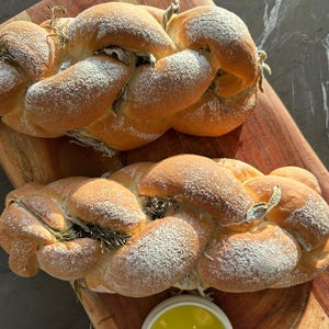 May include: Two braided loaves of bread dusted with powdered sugar and sprigs of rosemary, presented on a wooden cutting board. A small white bowl of olive oil is in the foreground.