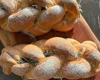 May include: Two braided loaves of bread dusted with powdered sugar and sprigs of rosemary, presented on a wooden cutting board. A small white bowl of olive oil is in the foreground.