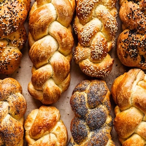 May include: Overhead shot of various braided loaves of bread. Some are sprinkled with sesame seeds, others with poppy seeds, and some are plain. The bread has a golden-brown crust and is arranged on a light-colored surface.