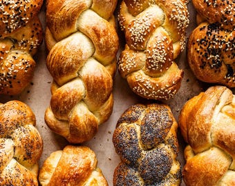 May include: Overhead shot of various braided loaves of bread. Some are sprinkled with sesame seeds, others with poppy seeds, and some are plain. The bread has a golden-brown crust and is arranged on a light-colored surface.