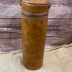 Peut inclure: Un porte-bouteille de vin cylindrique en cuir marron avec une poignée. Le porte-bouteille a une fermeture éclair et des coutures blanches. Le cuir a un aspect vieilli. Conçu pour contenir une bouteille de vin, environ 30 cm de hauteur.