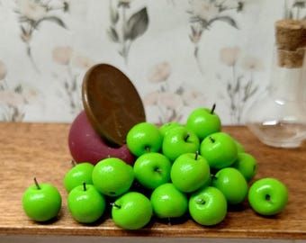 May include: A collection of miniature green apples with black stems, scattered on a wooden surface. A large brown coin and a dark red object are also present. A clear glass bottle with a cork stopper is in the background.