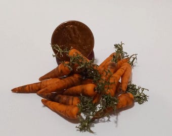 May include: A pile of miniature orange carrots with green leafy tops. A penny is partially visible in the background. The carrots are arranged on a white surface.