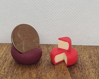 May include: Miniature red cheese wheels with a creamy interior, one partially sliced. A penny is nestled in a small, dark red holder. The items are on a wooden surface, suggesting a food-themed miniature.