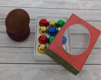 May include: A collection of small, colorful Christmas ornaments in a white box, with a red and green decorative box. A penny is placed on top of a dark purple object. The ornaments are red, gold, green, and blue.
