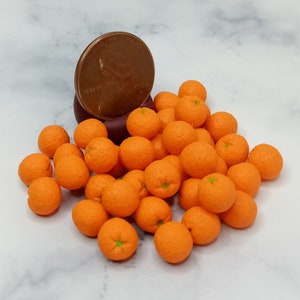 May include: A collection of miniature orange fruits with green stems, scattered on a surface. A copper penny is placed on a dark purple object, providing a size reference. The oranges are bright orange and appear to be made of a smooth material.