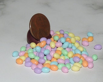 May include: A pile of pastel-colored candy-coated almonds, in shades of blue, green, yellow, pink, and orange, next to a copper penny. The candies are oval-shaped and scattered on a white surface.