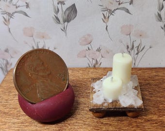 May include: A miniature dark red rounded holder containing a copper-colored US penny next to a small square wooden platform with four feet, topped with two off-white pillar candles surrounded by clear, jagged, translucent crystal-like fragments.