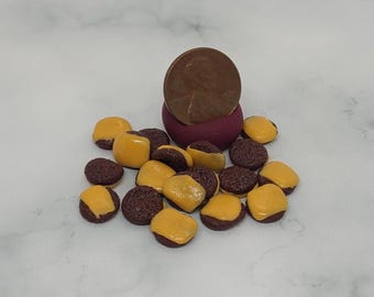 May include: Miniature food items, including small brown patties topped with yellow cheese, are scattered around a penny and a small, dark red container. The items resemble tiny cheeseburgers. The penny is a U.S. one-cent coin.