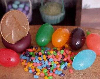 May include: A collection of colorful jelly beans in various sizes and colors, including green, orange, purple, red, and blue. A U.S. penny is next to a large purple jelly bean. Miniature baskets are in the background.